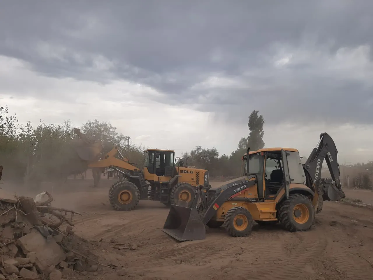 Pala cargadora y retroexcavadora trabajando en un terreno de tierra, con polvo y cielo nublado