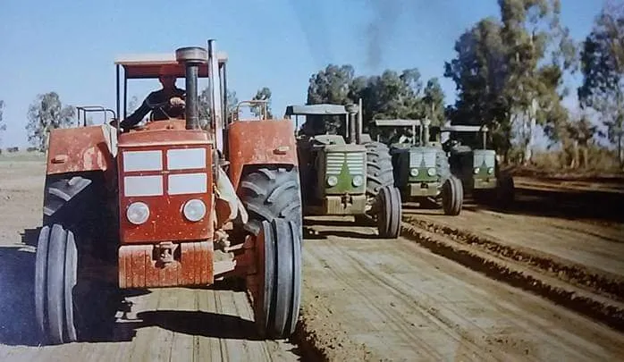 Tractor rojo con su operador al frente de una fila de tractores verdes sobre un camino de tierra