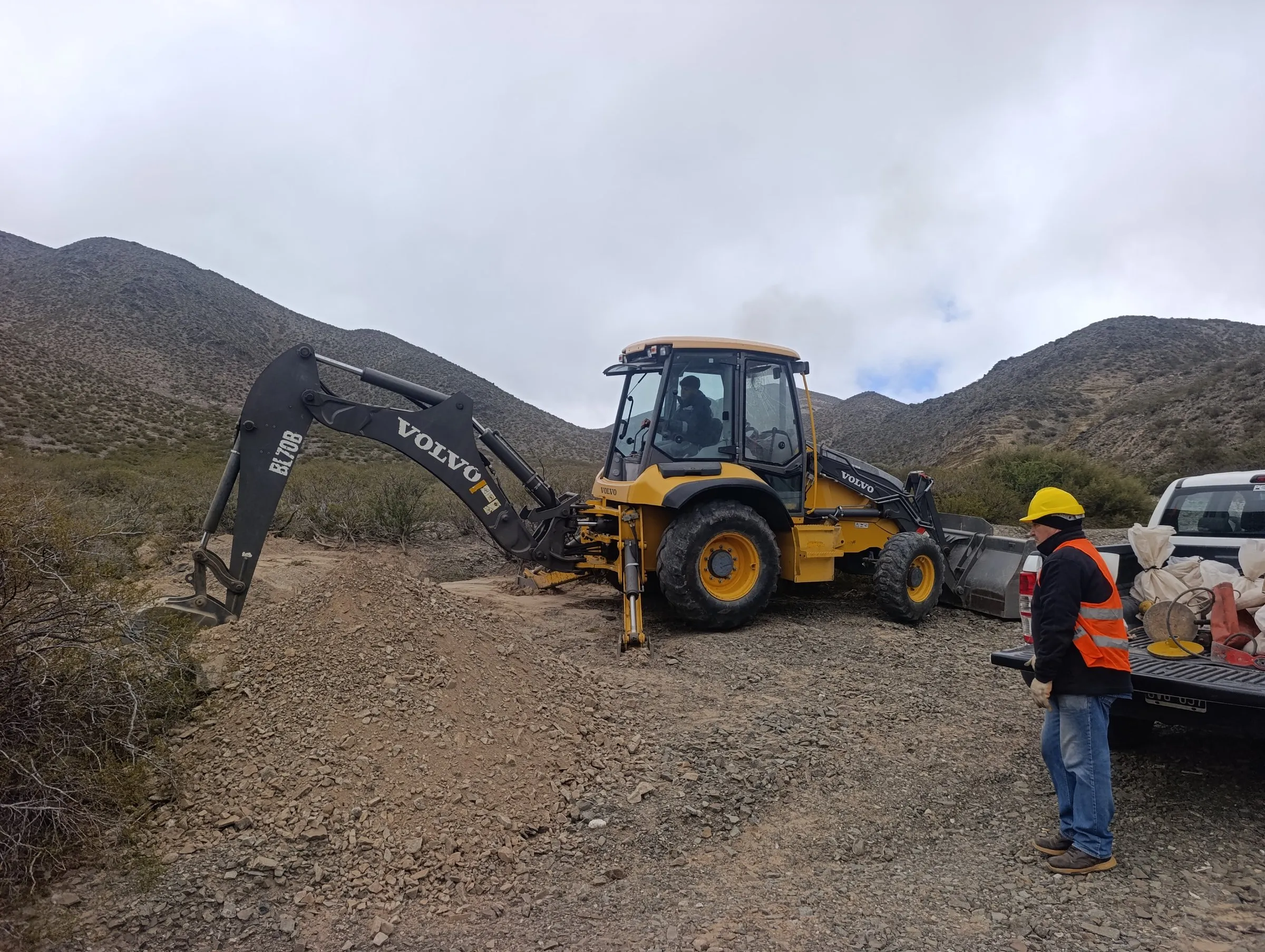 Operario con casco y chaleco reflectivo junto a una retroexcavadora Volvo trabajando en una zona de cerros